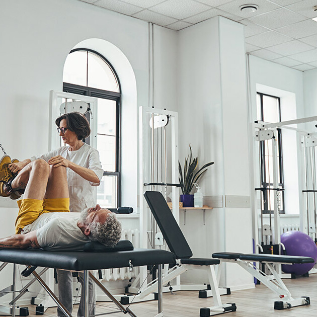 A physical therapist assists an older adult with leg exercises on a treatment table inside a bright rehabilitation clinic with strength and resistance equipment in the background.