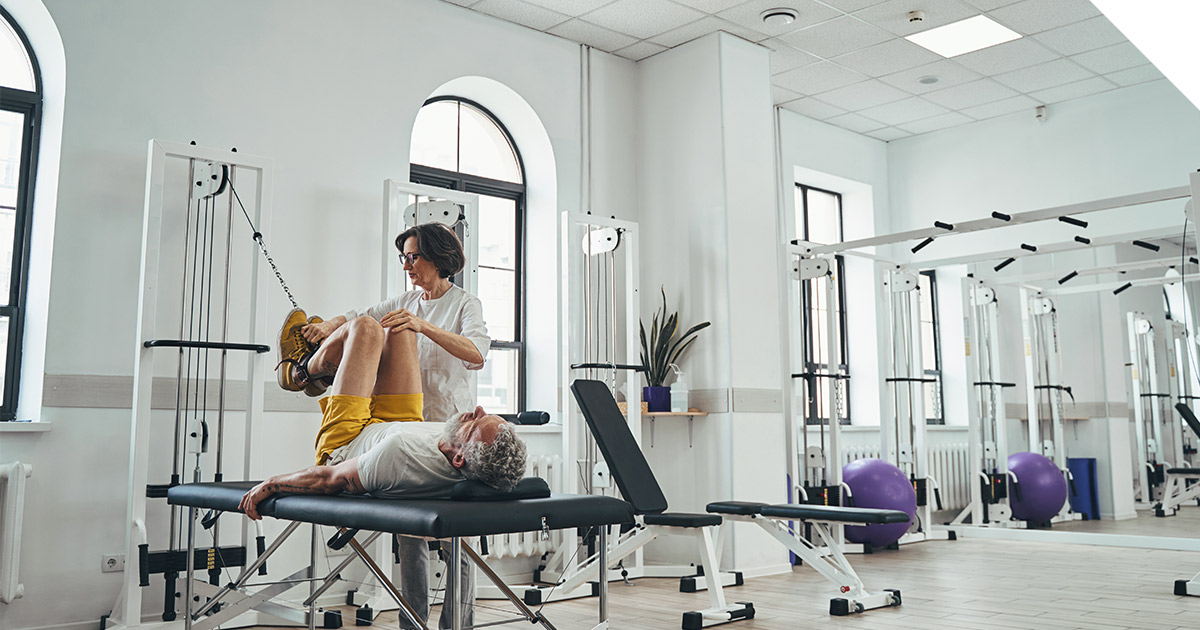 A physical therapist assists an older adult with leg exercises on a treatment table inside a bright rehabilitation clinic with strength and resistance equipment in the background.